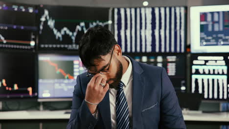 a businessman sits at a desk in front of multiple computer monitors displaying stock market data. he looks stressed and worried.