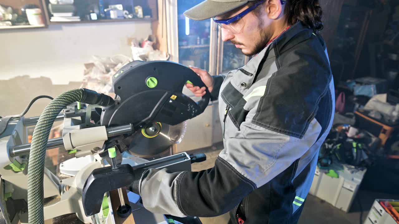 un joven con barba en un mono gris de profesión un carpintero trabaja con una máquina de corte circular en su taller de casa