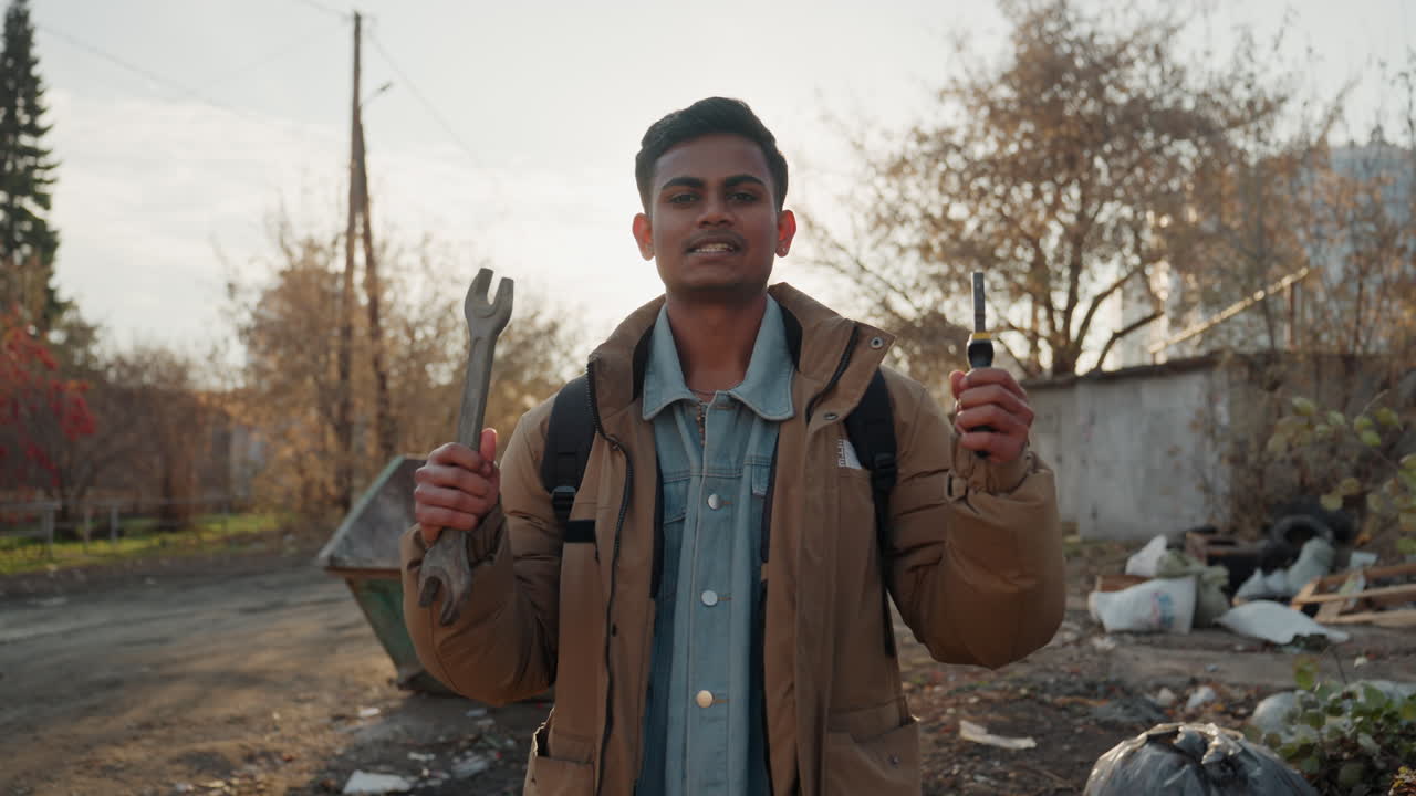 Homem sul-asiático segurando chave inglesa e chave de fenda perto de uma lixeira, sorrindo e confiante. Ferramentas reaproveitadas e uma pilha de lixo estão ao fundo, com luz solar contra a luz, transmitindo um espírito engenhoso de 'faz-tudo' com um tom esperançoso.