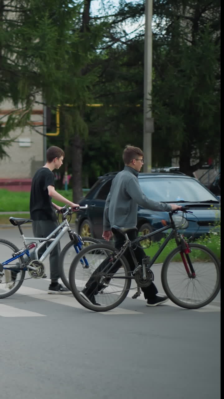 친구들이 자전거를 타고 보행자 교차로를 가로질러 나란히 걸어가고 있으며, 배경에는 두 사람이 걸어가고 있는 흐릿한 시야가 있고, 주차된 차가 보이고, 주변에는 나무가 보입니다.