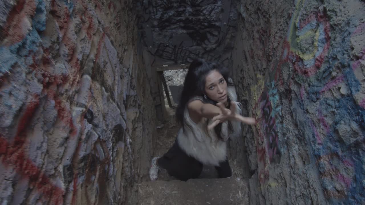 A dancer with long dark hair makes interactive hand gestures towards the camera in a gritty artistic performance