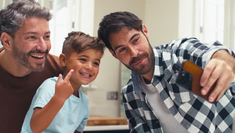 Same-Sex-Family-With-Two-Dads-Taking-Selfie-In-Kitchen-With-Son-Sitting-On-Counter