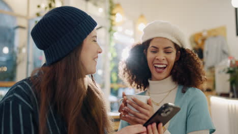 two friends laughing at something on a smartphone in a cafe