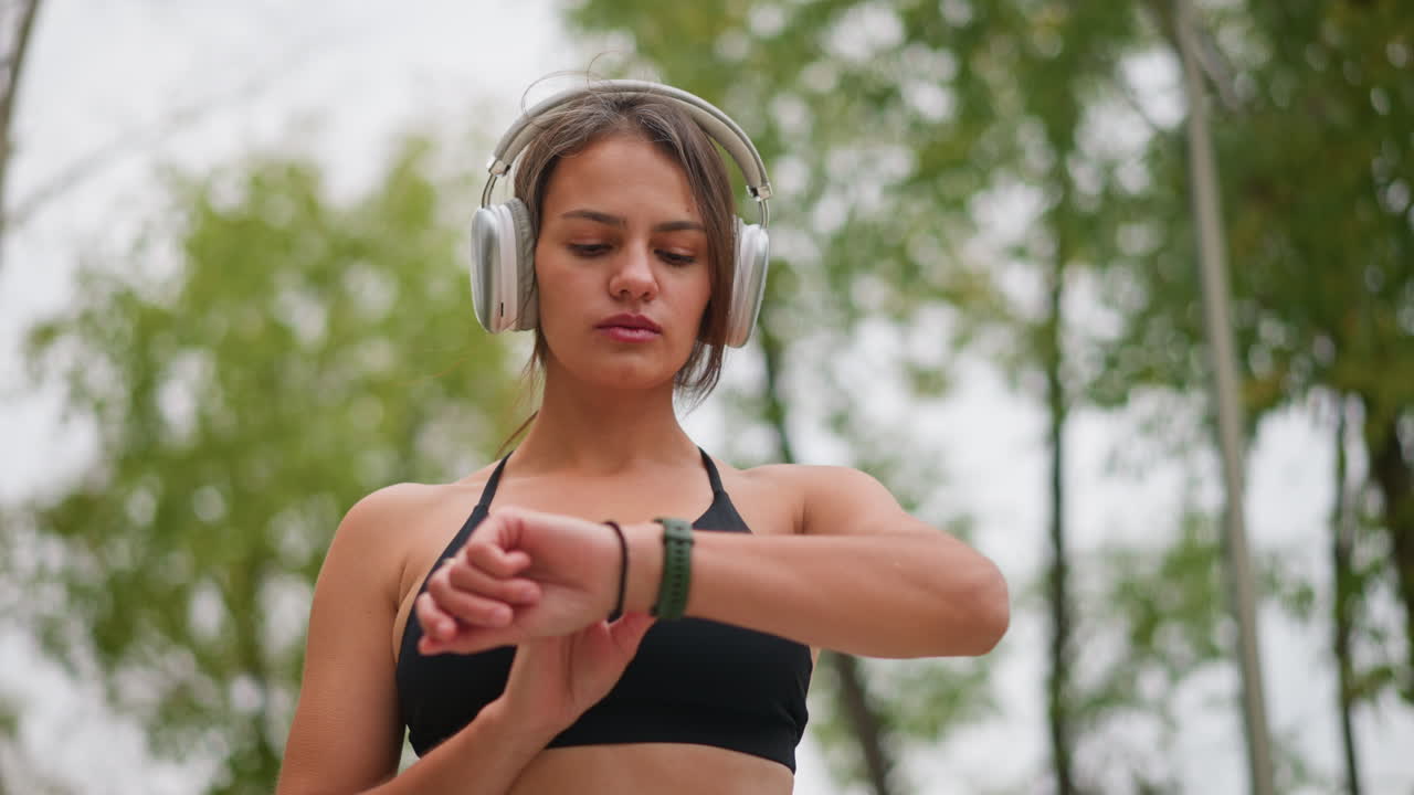 Close view of focused young lady standing outdoors, wearing headphones and checking time on her green wristwatch while black bangle on hand under bright natural setting