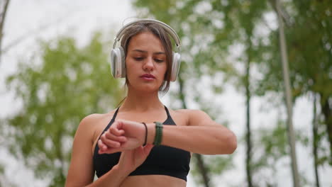 close view of focused young lady standing outdoors, wearing headphones and checking time on her green wristwatch while black bangle on hand under bright natural setting