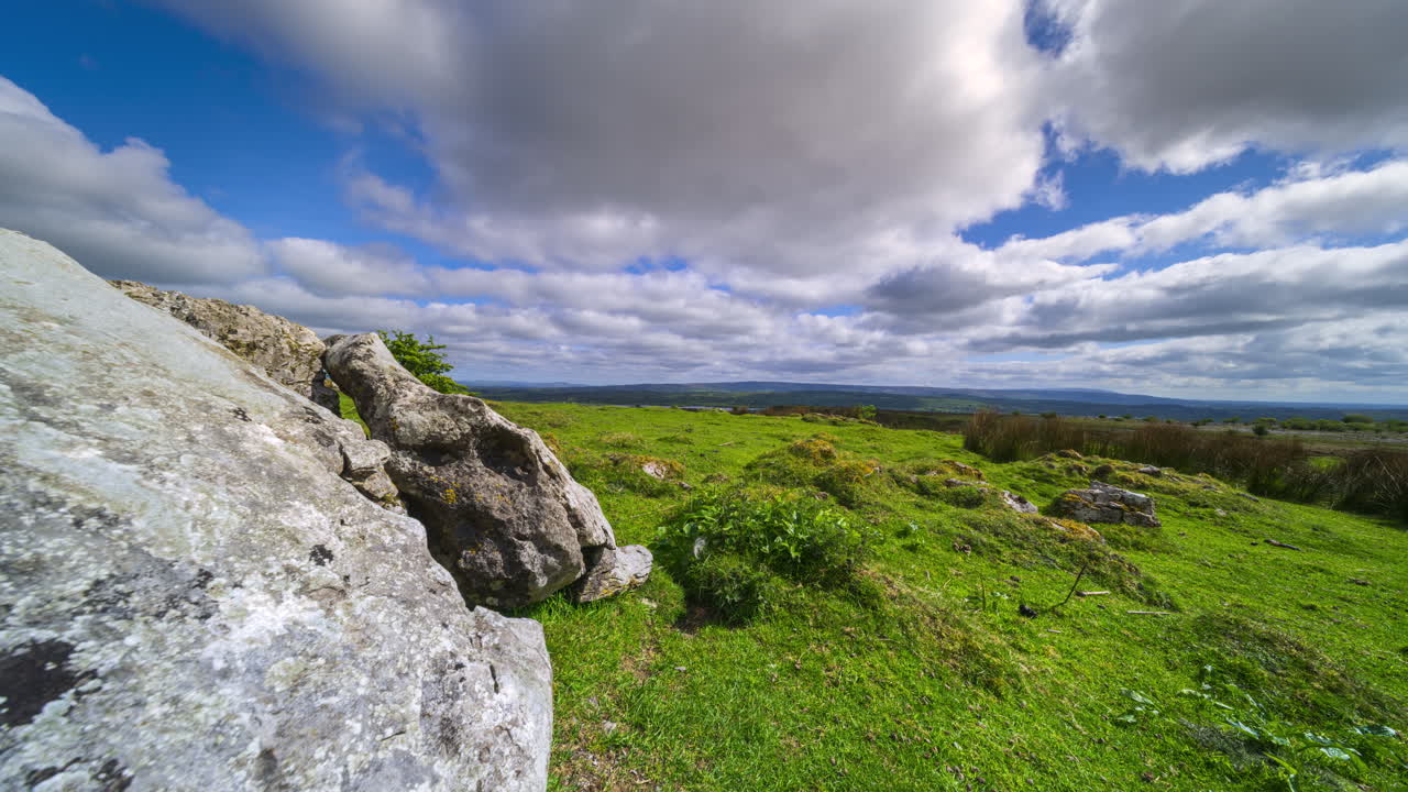 İrlanda, Sligo bölgesindeki Carrowkeel'den güneşli ve bulutlu bir günde çekilmiş, çimenlik ön planda kaya oluşumunun ve uzakta tepelerin bulunduğu kırsal manzaranın panoramik hareketli hızlandırılmış çekimi