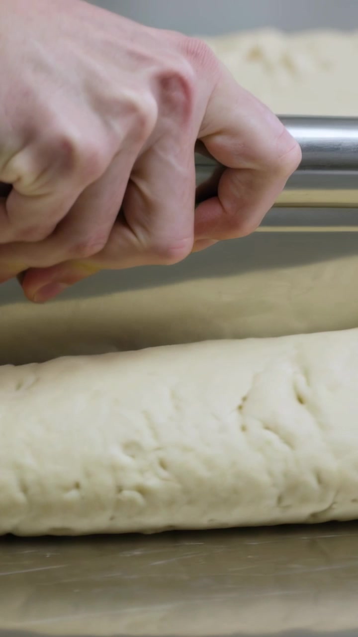 Vertical video: Pressing hands in white sleeve slicing loaf with bench scraper on steel for baking