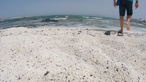 Hombre-Caminando-Descalzo-Hacia-La-Orilla-Del-Mar-En-La-Playa-De-Guijarros-Blancos-En-Fuerteventura,-Islas-Canarias,-España