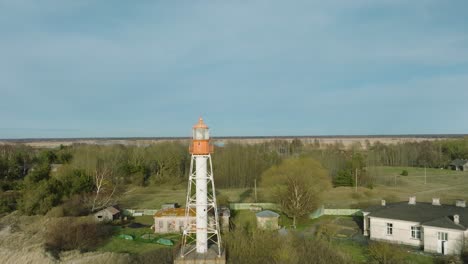 Aerial-establishing-view-of-white-colored-Pape-lighthouse,-Baltic-sea-coastline,-Latvia,-white-sand-beach,-large-waves-crashing,-sunny-day-with-clouds,-wide-drone-shot-ascending-backward