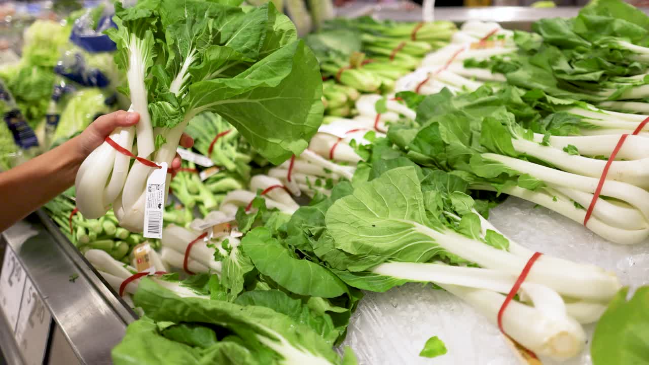 A person examines bok choy in a well-lit supermarket produce section. The scene captures fresh greens and careful selection