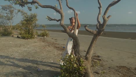 disparo en cámara lenta de una hermosa mujer en un vestido blanco flotante posando en una playa cerca de un árbol