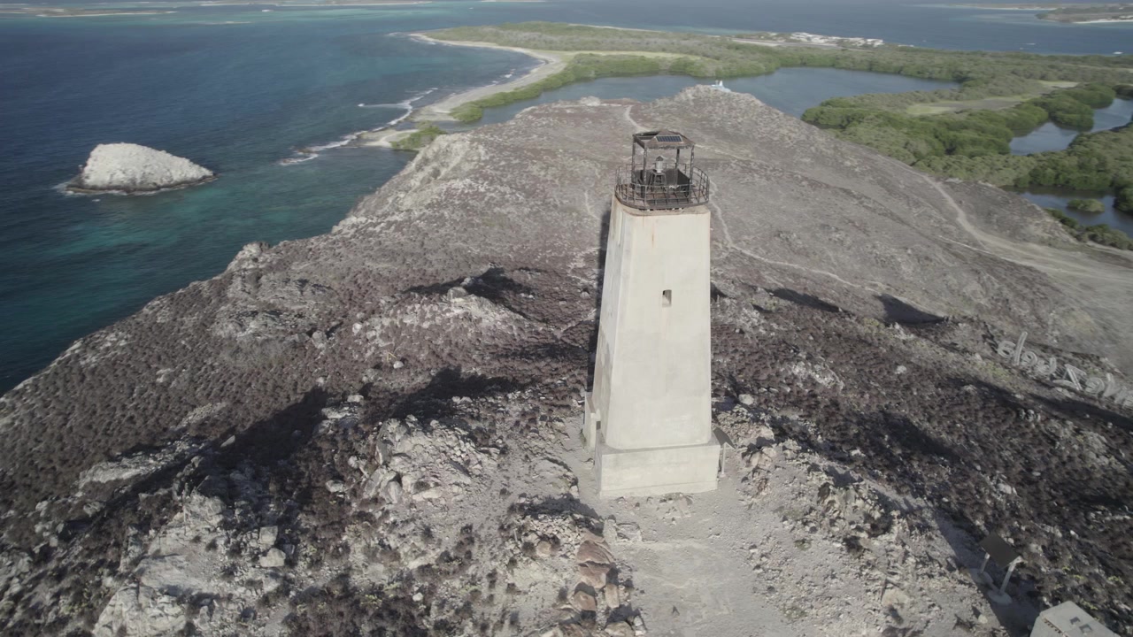 Drönarvy i omloppsbana över gammal fyr på stenig bergstopp nära kustlinjen och byn.
