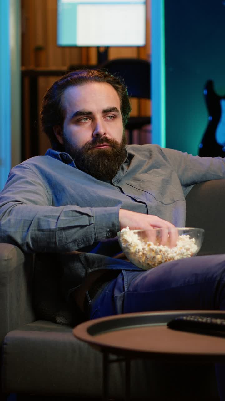 Vertikal video Huseier som kjeder seg hjemme mens han spiser snacks og ser på TV