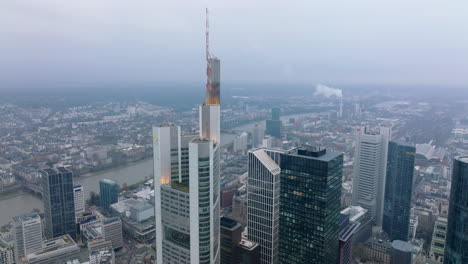 Erhöhte-Aufnahme-Von-Wolkenkratzern-In-Der-Innenstadt.-Fliegen-Sie-Um-Die-Spitze-Des-Modernen-Büroturms-Herum.-Stadt-Mit-Fluss-Im-Hintergrund.-Frankfurt-Am-Main,-Deutschland