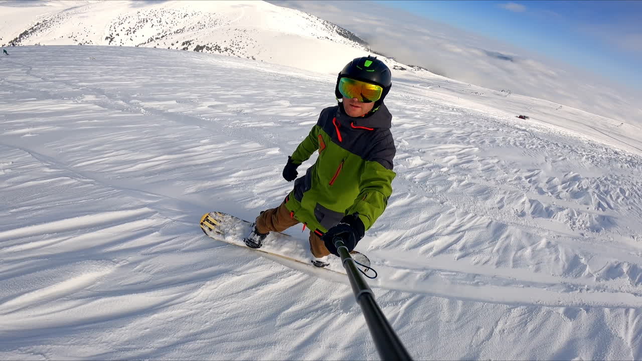 Snowboard descend by the mountain slope. Sportsman taking selfie of his ride and his fellow's ride. Stunning mountains at backdrop.