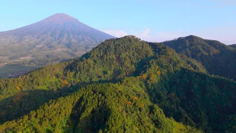 Mountain-slope-covered-in-dense-forest,-volcano-in-background,-aerial-drone-view