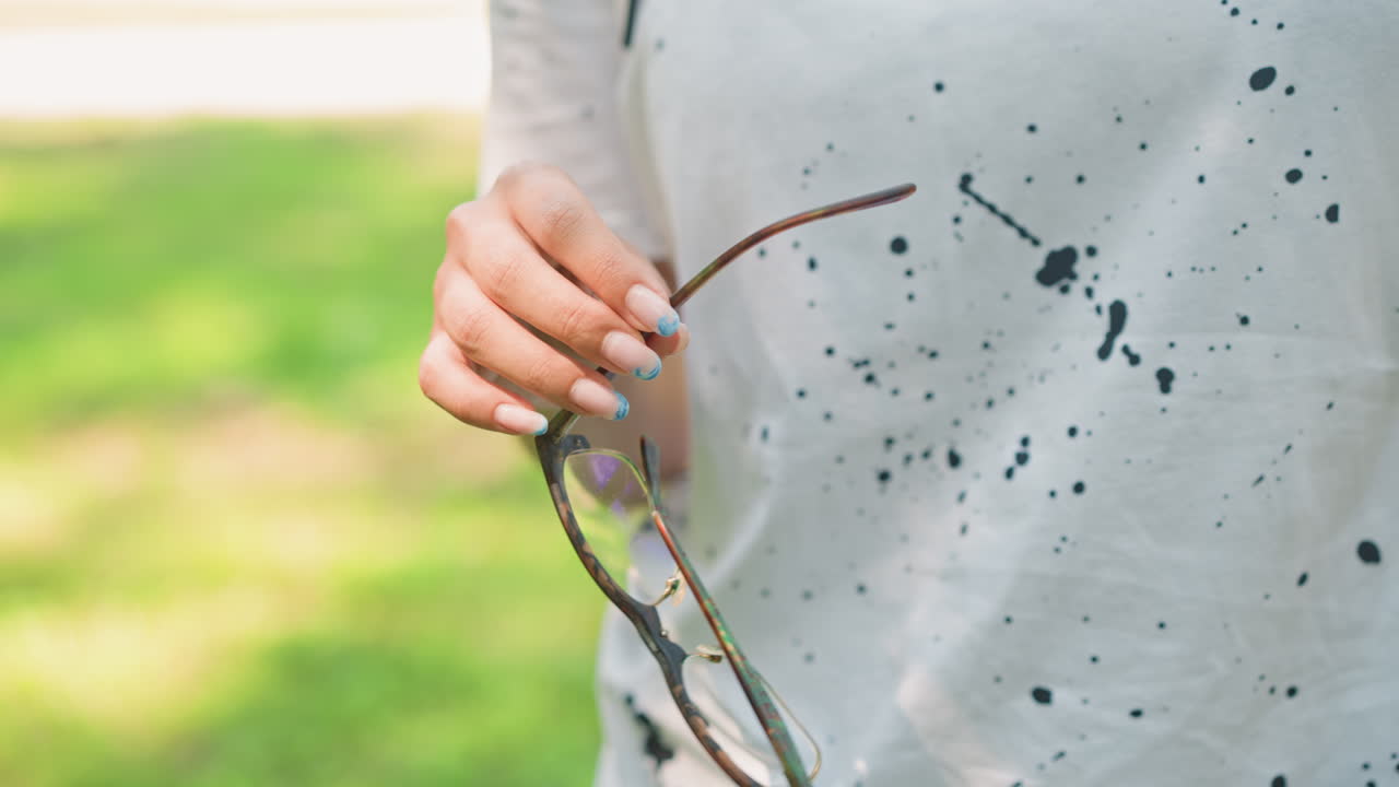 선글라스를 낀 백인 여성이 햇빛이 잘 드는 공원에서 손과 몸통을 클로즈업한 채 야외에 있어요. 부드러운 보케와 페인트 얼룩이 있는 캐주얼 티셔츠, 보이는 매니큐어, 부드러운 움켜쥠과 느린 동작이 느껴져요.