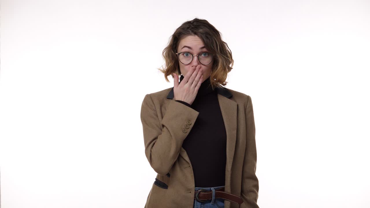 Curly haired european woman in stylish eyeglasses looking to the camera and showing silence gesture, keeping secrets. Metaphorically fastens her mouth and throws a key. Cheerfully smiling