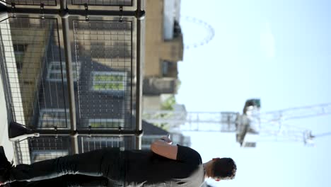 Man-using-a-smartphone-while-standing-on-a-rooftop-balcony-in-an-urban-setting-with-construction-equipment-and-a-Ferris-wheel-in-the-background-on-a-clear-day