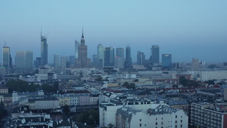 Backwards-fly-above-urban-neighbourhood.-Skyline-with-skyscrapers-in-background.-Aerial-shot-before-sunrise.-Warsaw,-Poland