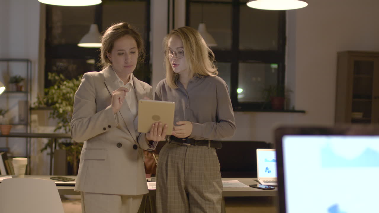 две женщины-коллеги разговаривают и смотрят планшет, стоя в офисе 1