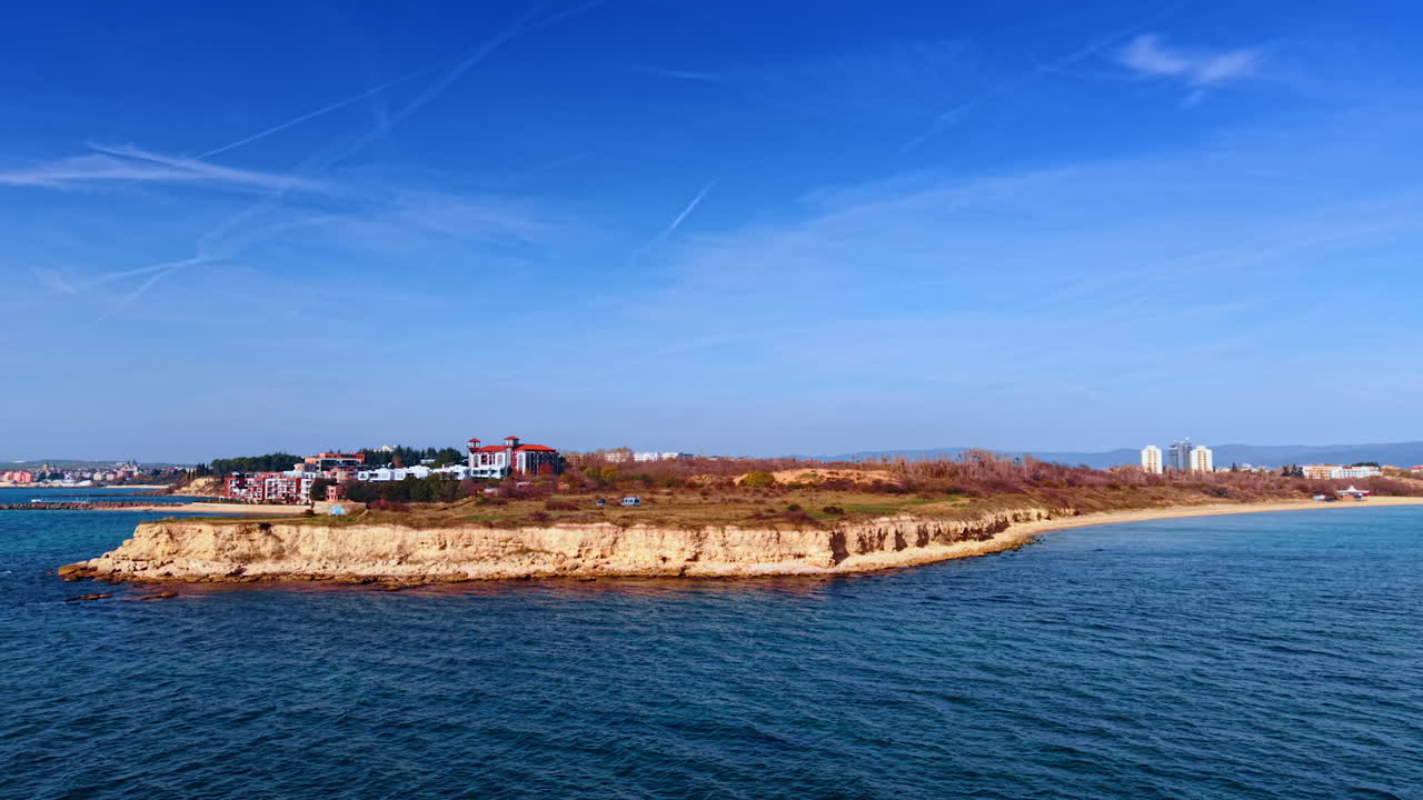 A small island stands in the water near a city. Buildings are visible on the island and along the coastline. The sky is clear with a few clouds. The view is taken during the day