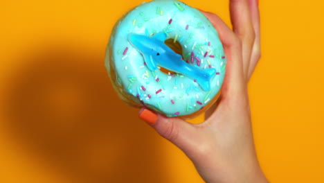 a child’s hand displays a playful blue frosted donut with rainbow sprinkles and a plastic shark figure on top, set against a bright yellow background.