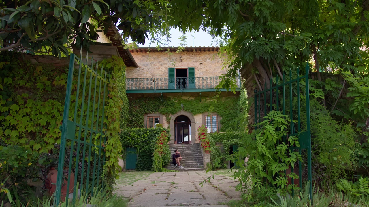 Inquadratura fissa di un uomo in nero che scende le scale di una villa toscana, sotto la luce del sole estivo, evocando mistero ed eleganza