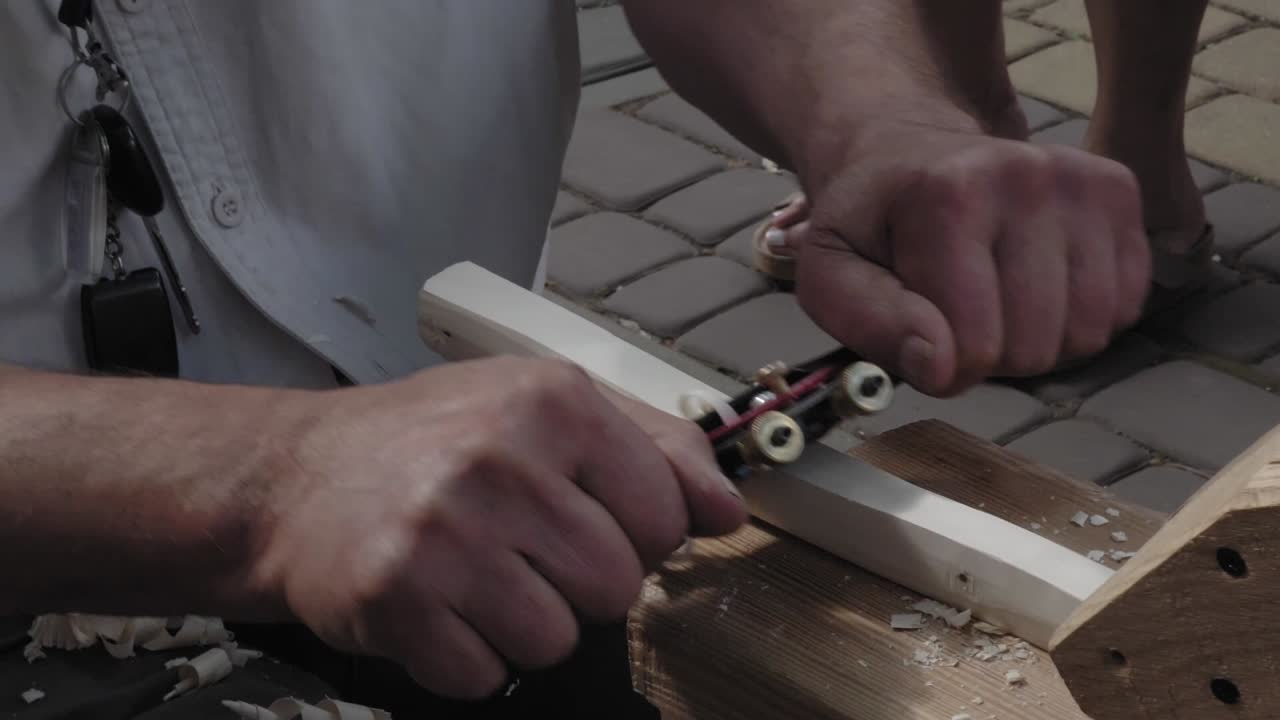 A traditional carpenter uses a metal planer to peel wood. He works outside during the event. People pass by.