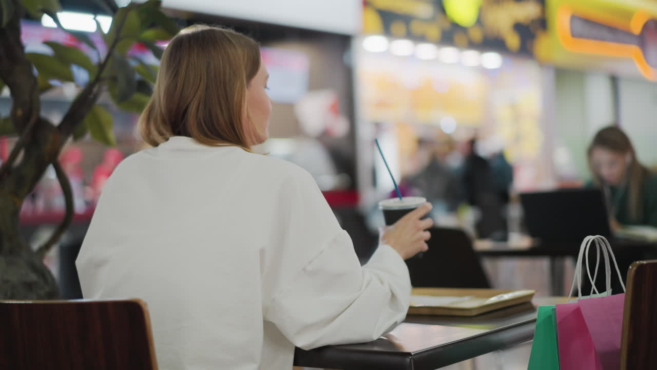 vista trasera de la dama en camisa blanca bebiendo café de la taza mientras está sentada en un restaurante dentro del centro comercial, el fondo incluye una vista borrosa de la gente