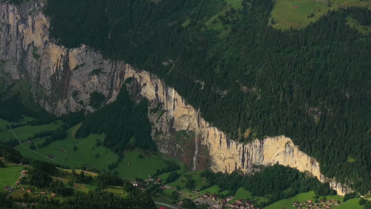 Aerial shot of dramatic cliffs and cascading waterfall in Lauterbrunnen Valley, Switzerland. The scene shows steep vertical rock walls, lush green meadows, and scattered alpine houses at the base