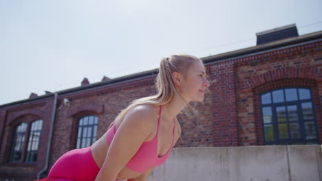 joven mujer rubia en ejercicios de color rosa con kettlebell por el edificio de ladrillos