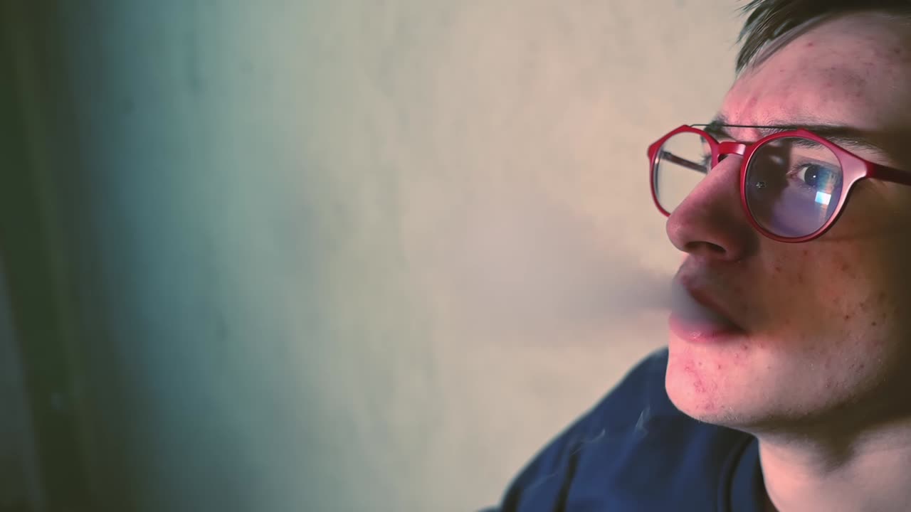Young man in red glasses vaping and exhales smoke indoor. Side view. Close up. Slow motion
