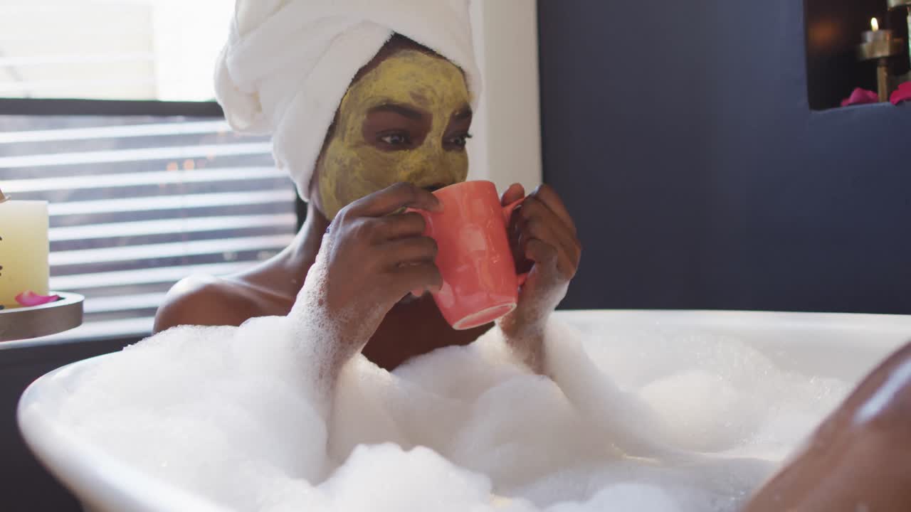 Smiling african american woman with towel and mask taking bath and drinking coffee in bathroom