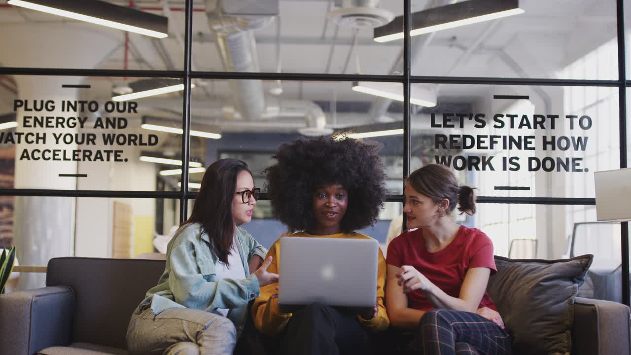 Three millennial women sitting on the sofa in an office lounge using a laptop together, front view