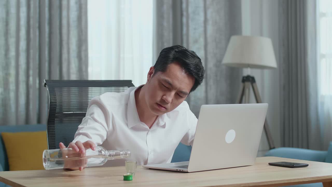 아시아인 남자가 술을 마시기 전에 글라스에 보드카를 부어서 에서 노트북으로 타이핑하고 있습니다.