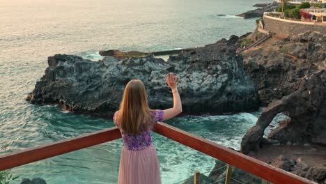 Young-Woman-Standing-on-Terrace-Balcony-Waving-Across-Coastal-Bay-Rocky-Cliffs-Along-Los-Gigantes-with-Turquoise-Ocean-Waves-in-the-Background-on-the-Canary-Islands,-Tenerife