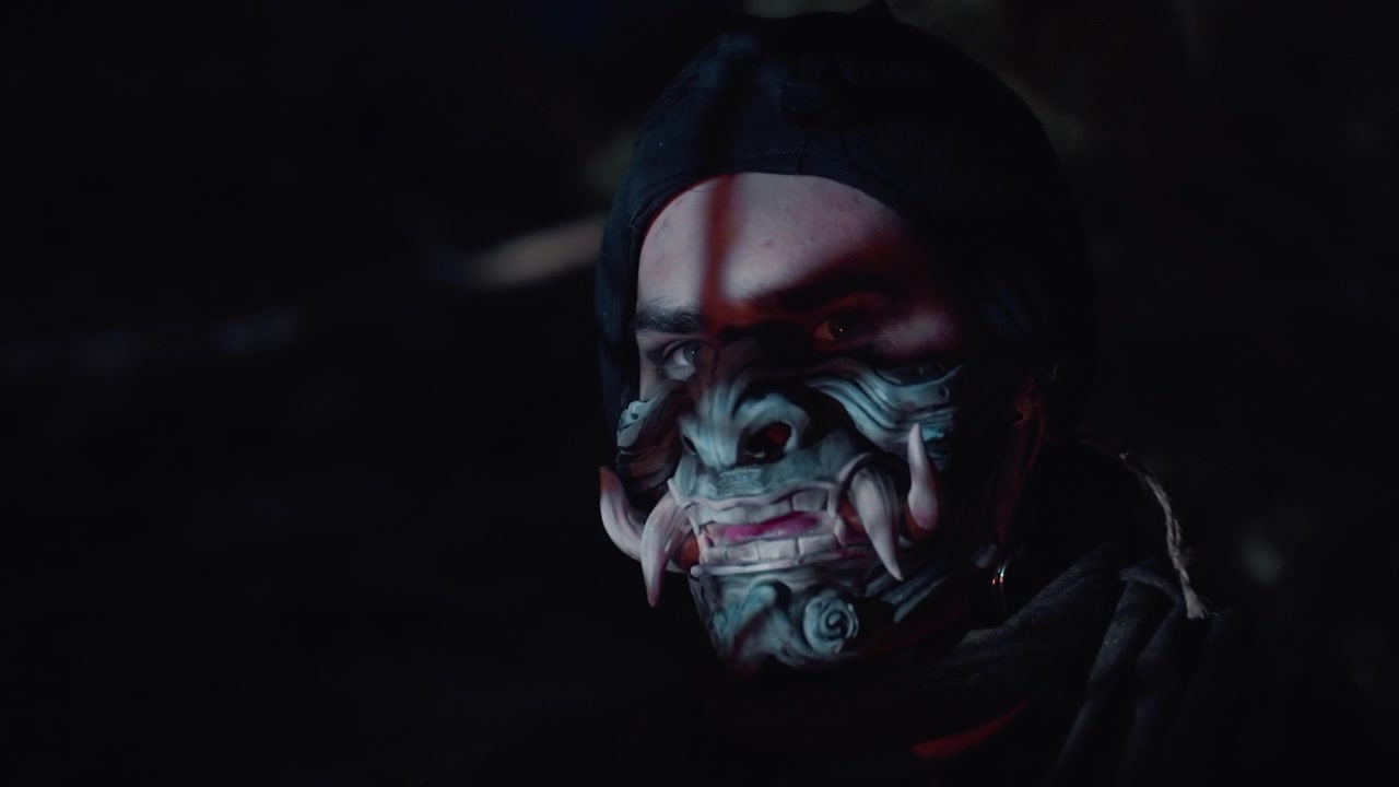 Close-up nighttime portrait of person wearing decorative patterned face mask, head slightly turned and tilted, eyes open and focused, subtle forward lean suggesting careful movement in dark moody