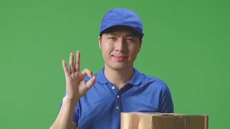 푸른 유니폼을 입은 아시아 남성 택배사의 클로즈업은 스튜디오에서 녹색 스크린 배경에 상자를 배달하는 동안 은 몸짓과 미소를 짓고 있습니다.