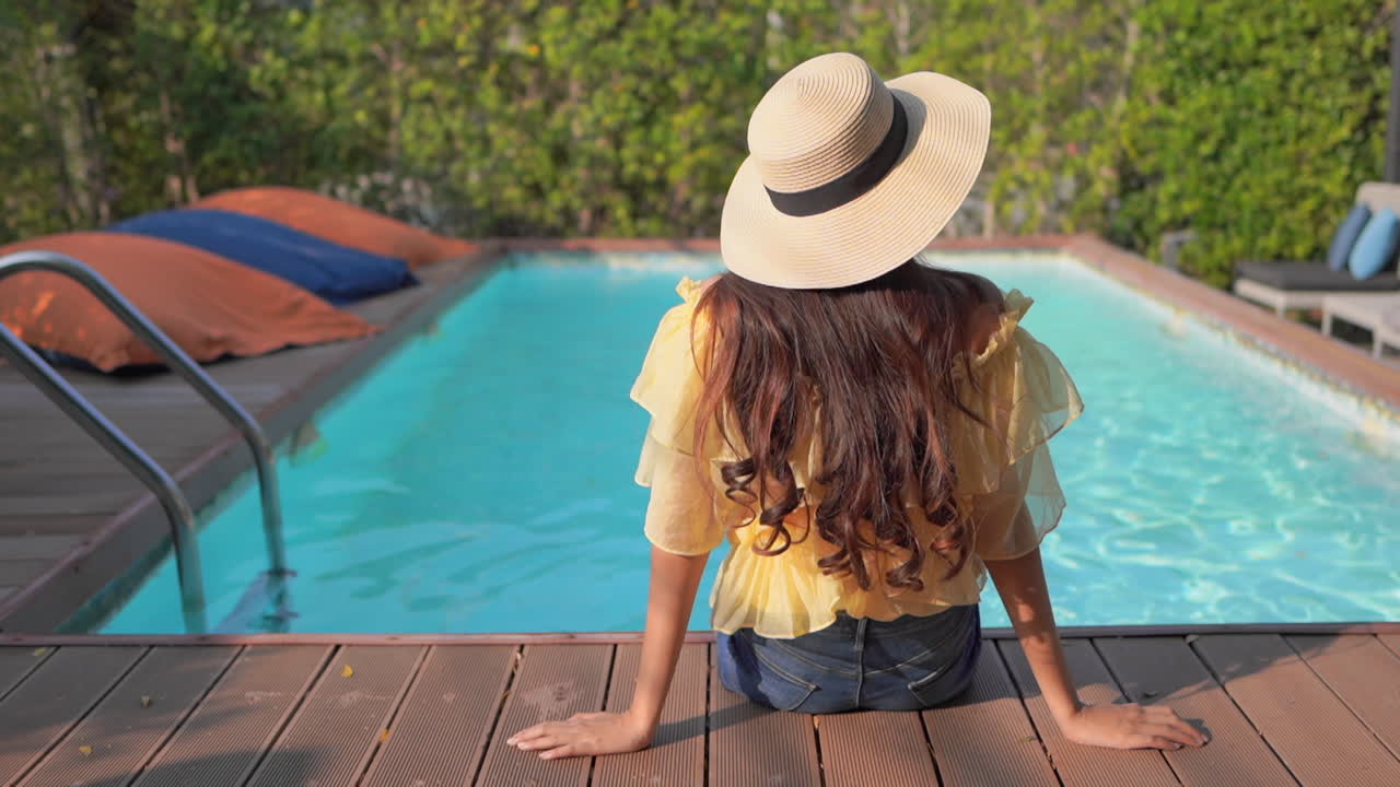 Rückenansicht einer weiblichen Touristin, die an einem Swimmingpool sitzt, einen Sonnenhut trägt und sich in der Poolvilla umsieht.
