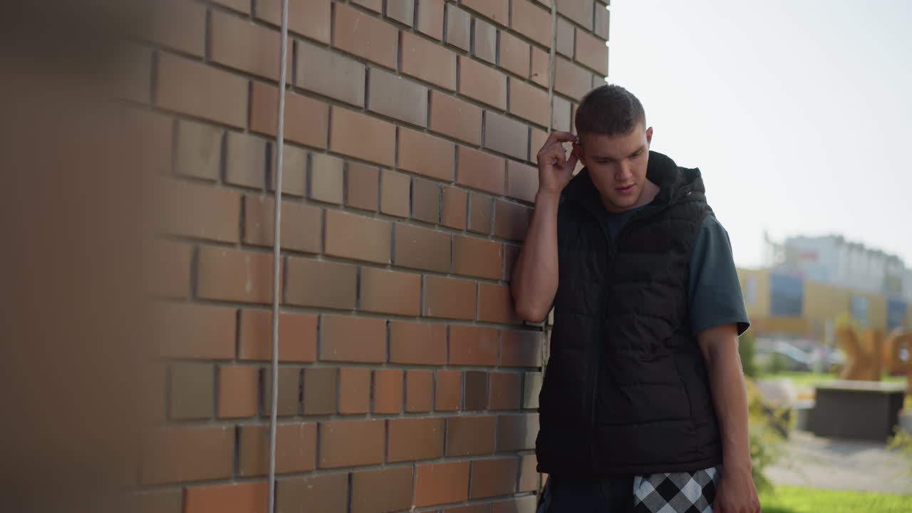 student walks up to brick building and leans against it while pulling cigarette from ear and preparing to light it with lighter, wearing dark vest and plaid shirt tied around waist
