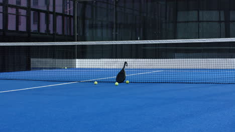 Padel tennis racket and balls on blue indoor court near net, ready for play, copy space