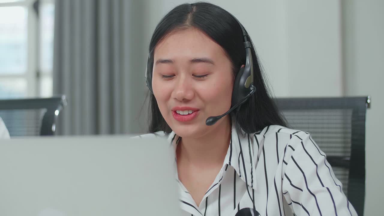 Front View Of An Asian Woman Call Center Agent Wearing Headset Typing On The Computer While Speaking To Customer On The Call During Working With Colleagues At The Office