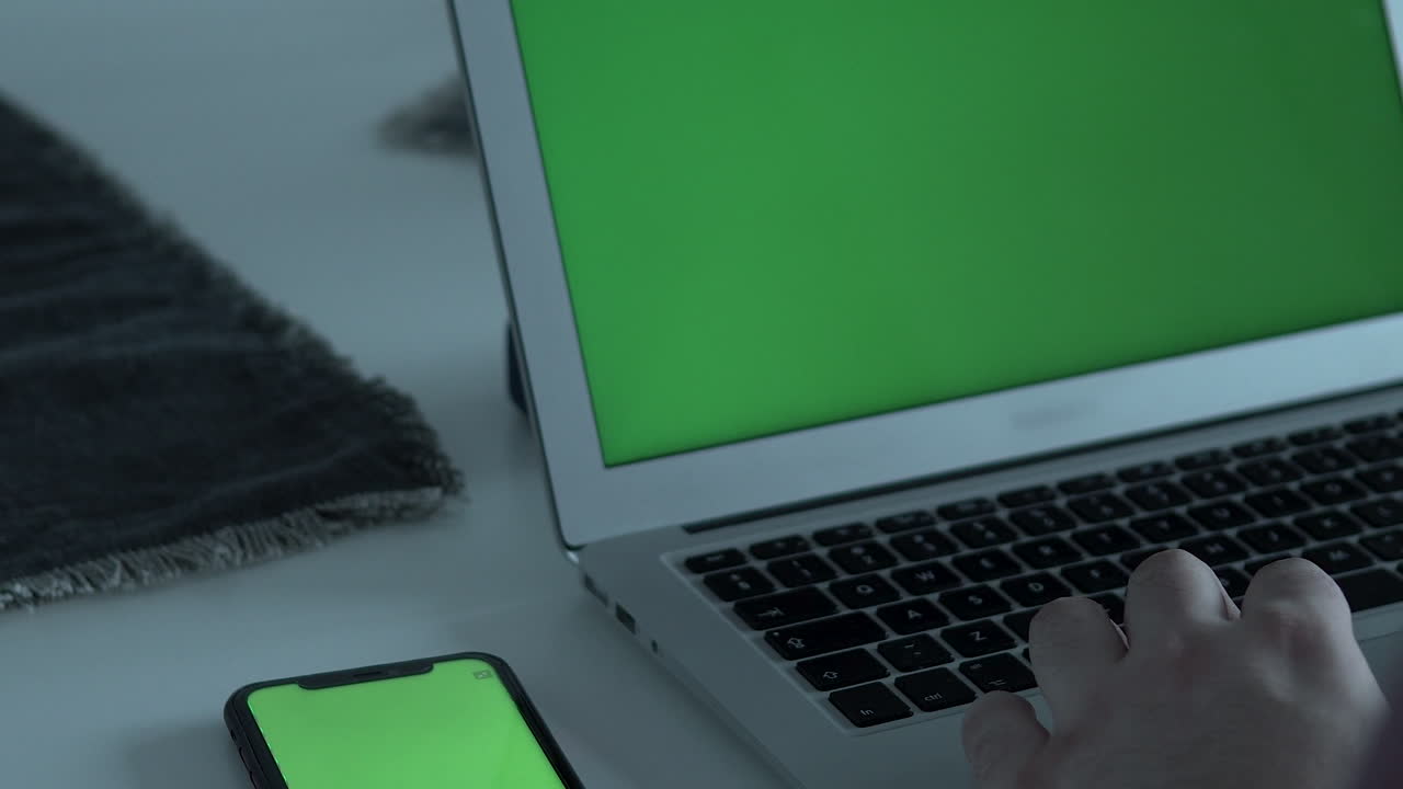Primer plano de un hombre tecleando en el teclado de su portátil; junto a él, está su teléfono móvil, pantalla verde.