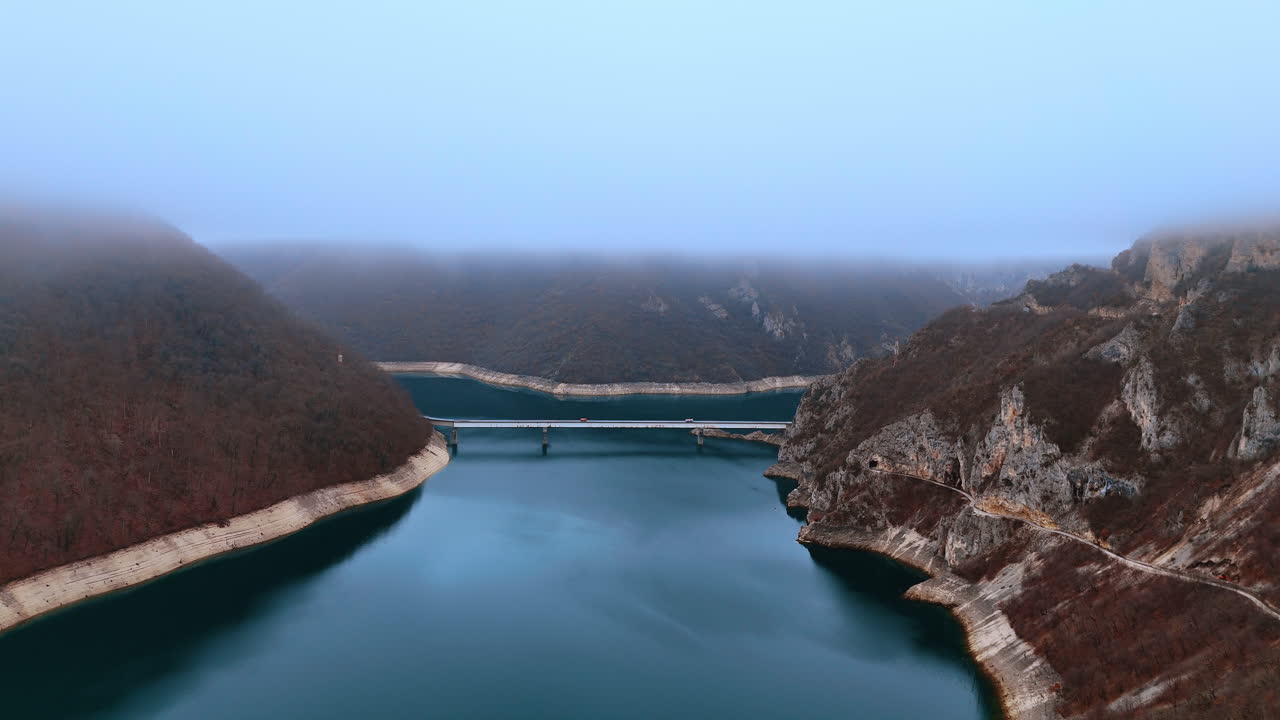 Piva lake bridge crossing turquoise water under foggy sky. Aerial view of a concrete bridge over Piva reservoir surrounded by steep hills and dense low mist