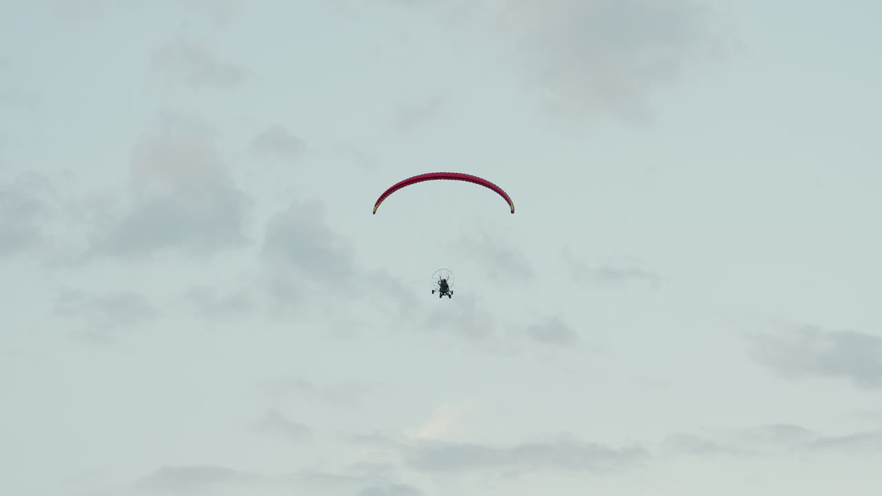 back view of paraglider pilot flying high under red canopy in cloudy sky with motorized frame visible capturing serene aerial adventure and open air exploration above misty horizon