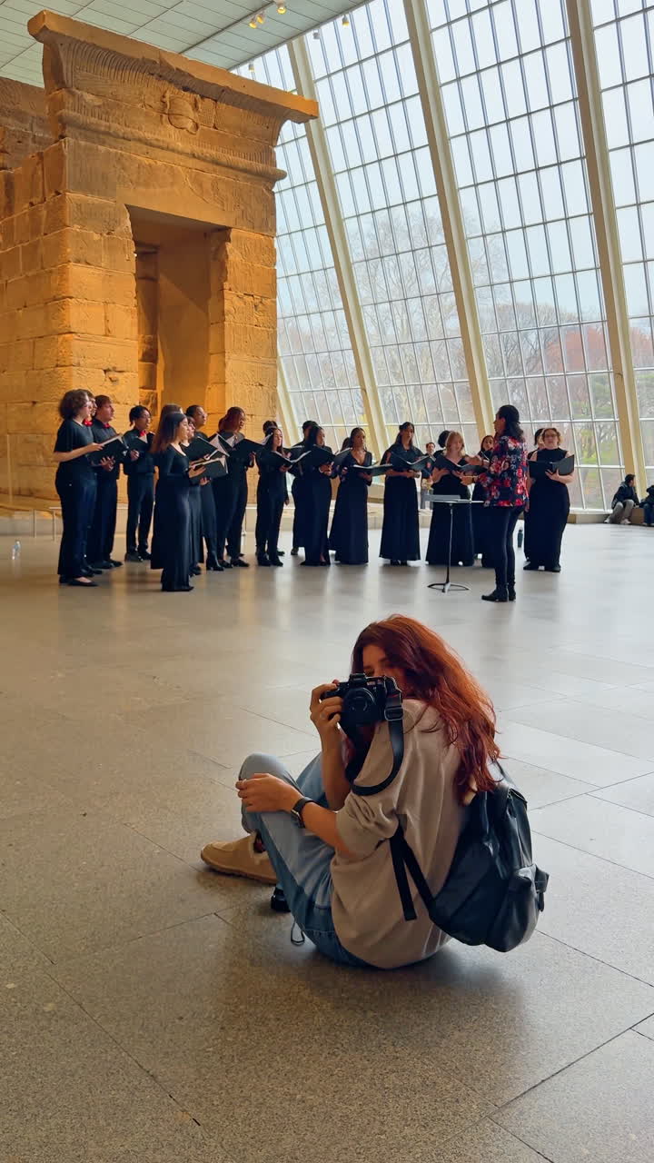 New York, AS, 2 Maret 2026: Seseorang menonton pertunjukan paduan suara di depan dinding kaca besar. Sudut lebar penonton yang duduk di lantai mengamati ansambel paduan suara di aula museum berdinding kaca di New York.