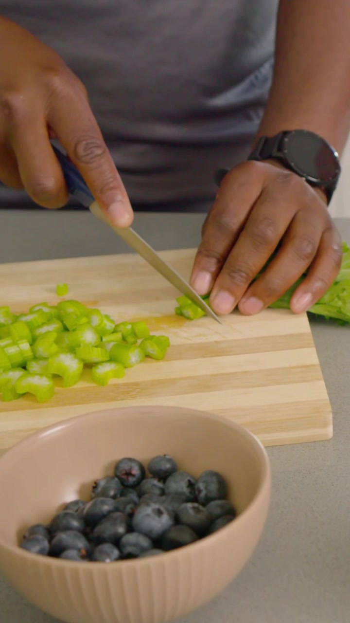 Video verticale: Posizionamento di un gambo, mani che affettano con un coltello blu su un tagliere sul bancone per insalata e frutti di bosco