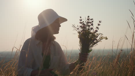 woman in white hat sitting in golden grassland gazing thoughtfully at bouquet of wildflowers in hand under warm sunset light, peaceful expression, serene outdoor nature scene with glowing