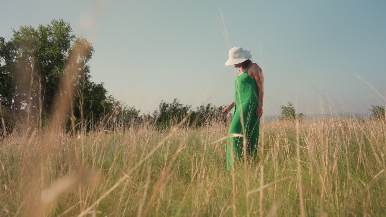 woman in green dress stands among tall golden grass with hand behind back, gently pulling single grass blade, sun shining on relaxed figure under soft blue sky